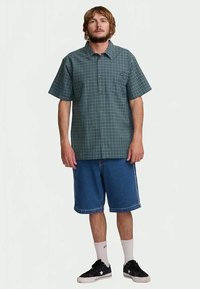 Man staat in een groen geruit kortebloezenshirt, blauwe denim short, witte sokken en zwarte sneakers op een effen achtergrond.