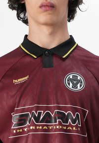 Bordo spalvos polo marškiniai su juodu apykakle, turintys geltonus akcentus, logotipą ant krūtinės ir raštuotą audinį. Tekste yra "hummel" ir "SWARM."