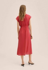 Femme aux cheveux blonds ondulés portant une robe midi corail sans manches cintrée à la taille et des sandales à talons kitten noires, se tenant de dos.