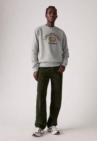 Man in een grijze Levi's Athletics sweatshirt, donkergroene corduroybroek en zilverwitte sneakers, staand tegen een lichte effen achtergrond.