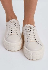 Beige leren sneakers met platte veters, verhoogde rubberen zolen met een gemarmerd patroon, en subtiele blauwe stikselaccenten.