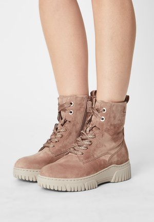 Bottines en daim brun clair avec semelles épaisses beiges, lacets à l'avant, fermeture éclair latérale, portées sur des jambes nues contre un fond uni.