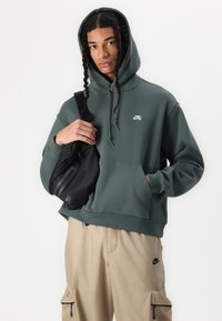 Zelena kapuca s prednjim žepom in majhnim belim logotipom, v kombinaciji s bež cargo hlačami in črno torbico preko telesa.