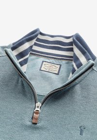 Pull-over bleu clair à fermeture éclair quart de tour avec un tissu texturé, un col rayé bleu marine, une fermeture éclair argentée et un petit logo sur le devant.