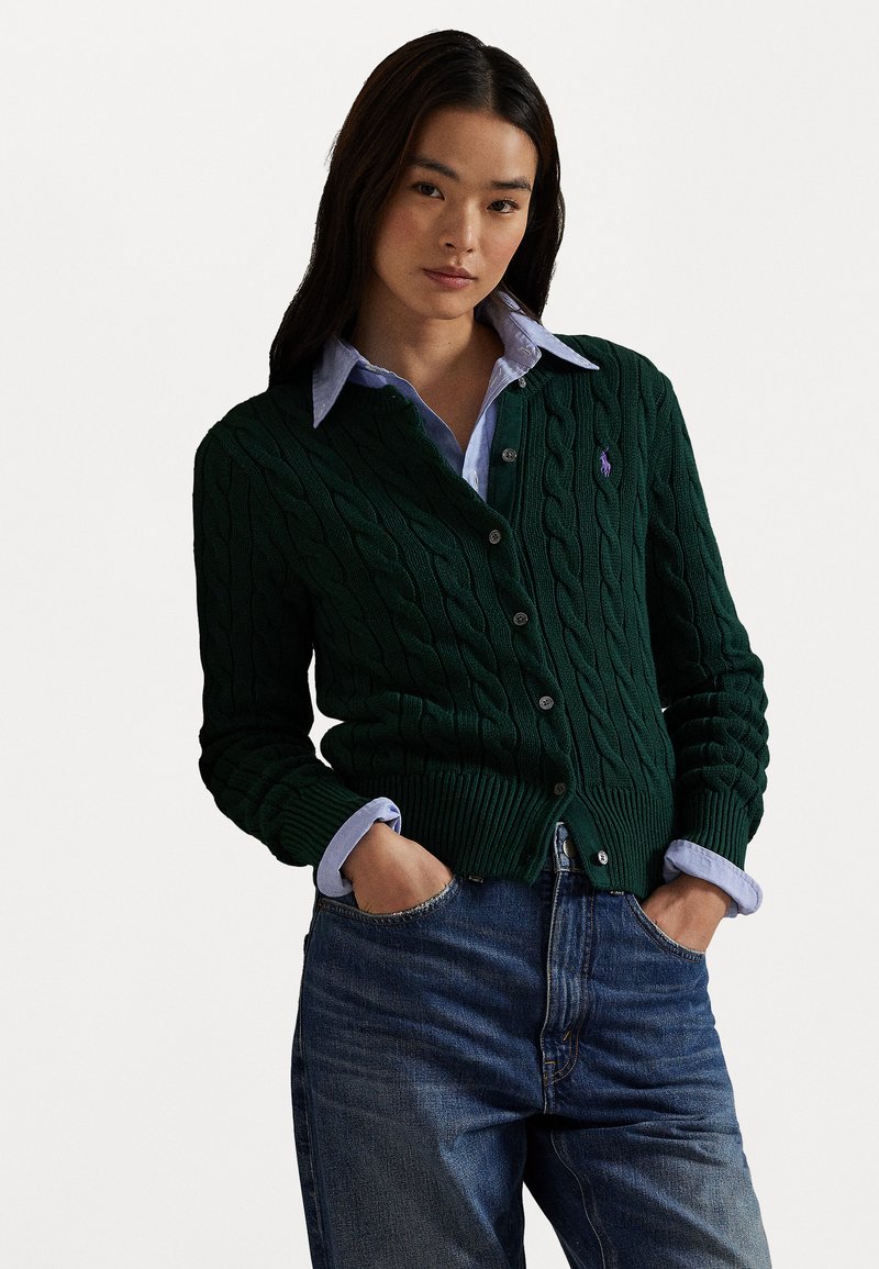 Grøn kabelstrik cardigan med knaplukning og ribbet kant. Båret over en lys blå skjorte med krave, parret med blå jeans.