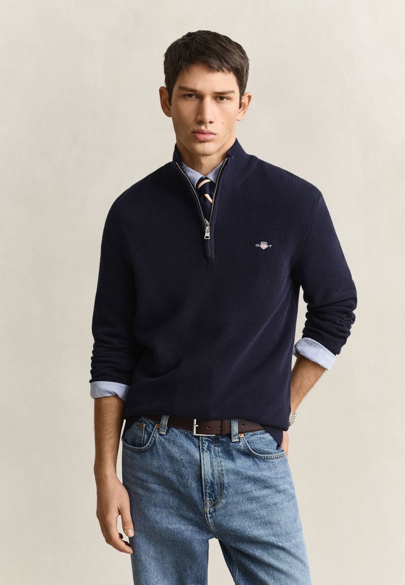 Jeune homme portant un pull bleu marine à demi-zip sur une chemise bleue et une cravate rayée, avec un jean bleu clair et une ceinture marron, debout devant un fond uni.