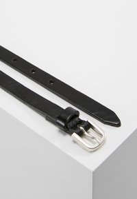 Černý kožený pásek se stříbrnou sponou, obsahující otvory pro úpravu velikosti, hladký povrch, zúžené konce a minimalistický design.