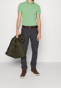 Grön polo skjorta, mörkgrå cargo-byxor, brun bälte, bruna snörskor. Håller en mörkgrön dunjacka med dragkedjor och huva.