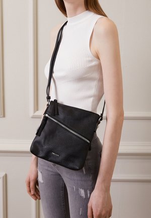 Woman wearing sleeveless white top and gray ripped jeans with a black crossbody bag featuring a diagonal zipper and adjustable strap.
