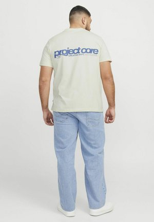 Lichtbeige katoenen t-shirt met het "project core" logo in het blauw op de achterkant, gecombineerd met lichtblauwe denim jeans en witte sneakers.