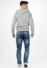 Felpa grigia con tasca a marsupio, abbinata a jeans blu consumati con cuciture visibili e cinque tasche. Dettagli di sneakers bianche.