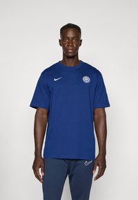 Sötétkék pamut póló kerek nyakkal, rövid ujjakkal, fehér Nike logóval a mellkason és egy kör alakú emblémával.