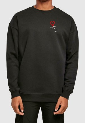 Person trägt einen schwarzen Pullover mit einem kleinen roten tropfenden Herz und weißem Text auf der oberen linken Brust, schwarze Hose sichtbar.