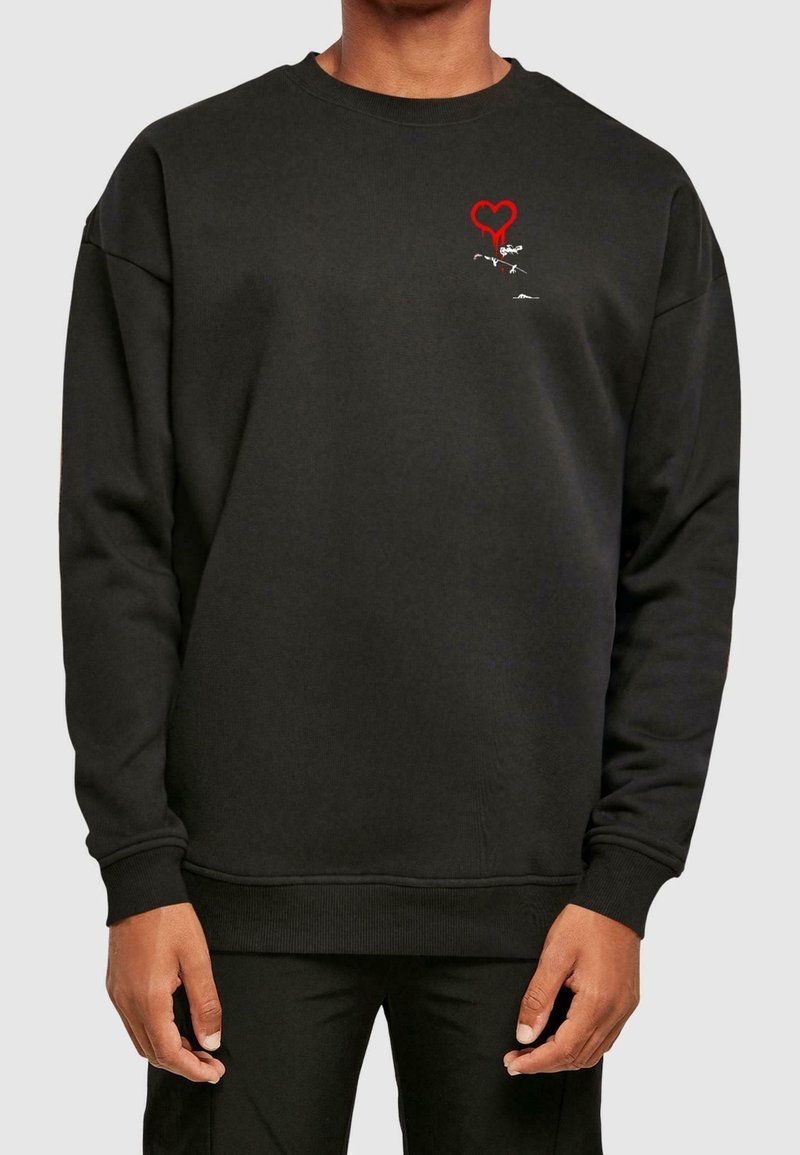 Personne portant un sweat-shirt noir avec un petit cœur rouge qui dégouline et un texte blanc sur le haut gauche de la poitrine, pantalon noir visible.