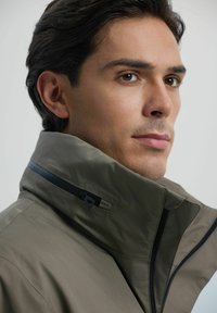 Lekki oliwkowy zielony kurtka z wysokim kołnierzem, czarnymi akcentami i gładką teksturą. Posiada zamek błyskawiczny i szczegół z trójkątnym metalowym elementem.