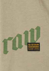 Beige fabric with green Gothic-style text "raw" and a black label patch reading "G-STAR ORIGINALS RAW DENIM" in yellow and orange.
