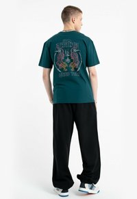 Młody mężczyzna ubrany w turkusową koszulkę z grafiką Muay Thai, czarne luźne spodnie i sneakersy, stoi tyłem z rękami za plecami.