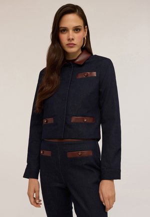 Veste en denim foncé coupe cropped avec des accents en cuir marron, dotée d'un col, de fermetures à boutons et de poches à l'avant. Texture lisse, coupe ajustée.