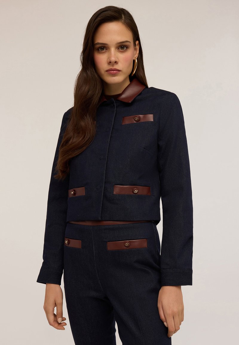 Dunkelblaue Jeansjacke im Cropped-Design mit braunen Lederakzenten, einer kragenförmigen Halslinie, Knopfverschluss und vorderen Taschen. Glatte Textur, taillierte Passform.