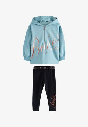 Junge Person trägt einen türkisfarbenen, übergroßen Hoodie mit dem Schriftzug "Baker", schwarze Leggings mit dem gleichen Schriftzug und weiße Sneaker und steht vor einer schlichten Wand.