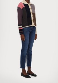 Multikleurige gebreide cardigan met crèmekleurige kraag, knoopsluiting en verschillende textuurpatronen; gecombineerd met blauwe jeans en zwarte platte schoenen.