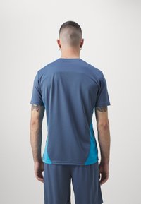 Tumši zils īsās piedurknes sporta krekls ar tirkīza sānu paneļiem, izgatavots no teksturēta auduma. Iezīmējas ar apaļu kakla izgriezumu un kontrastējošu šuvēm.