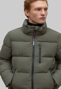 Chaqueta acolchada en verde oliva, con un alto cuello, diseño de acolchado horizontal, cremallera frontal y un bolsillo en el pecho con cremallera y acentos en contraste.