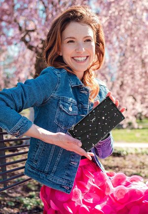 Black zippered wallet with pink and blue polka dots, quilted design. A woman in a denim jacket holds it against a backdrop of flowers.