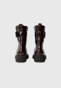 Brown patent leather ankle boots with a chunky sole, side zipper closure, and a strap with a buckle detail. Smooth texture and glossy finish.