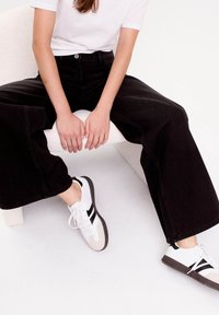 Sorte vide ben jeans og hvide sneakers med grå detaljer. Jeansene har høj talje, mens sneakersene har kontrasterende sorte striber og snørebånd.