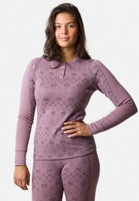 Ung kvinde iført matchende pink mønstrede termiske top og leggings, stående med den ene hånd på hoften mod en ensfarvet baggrund.