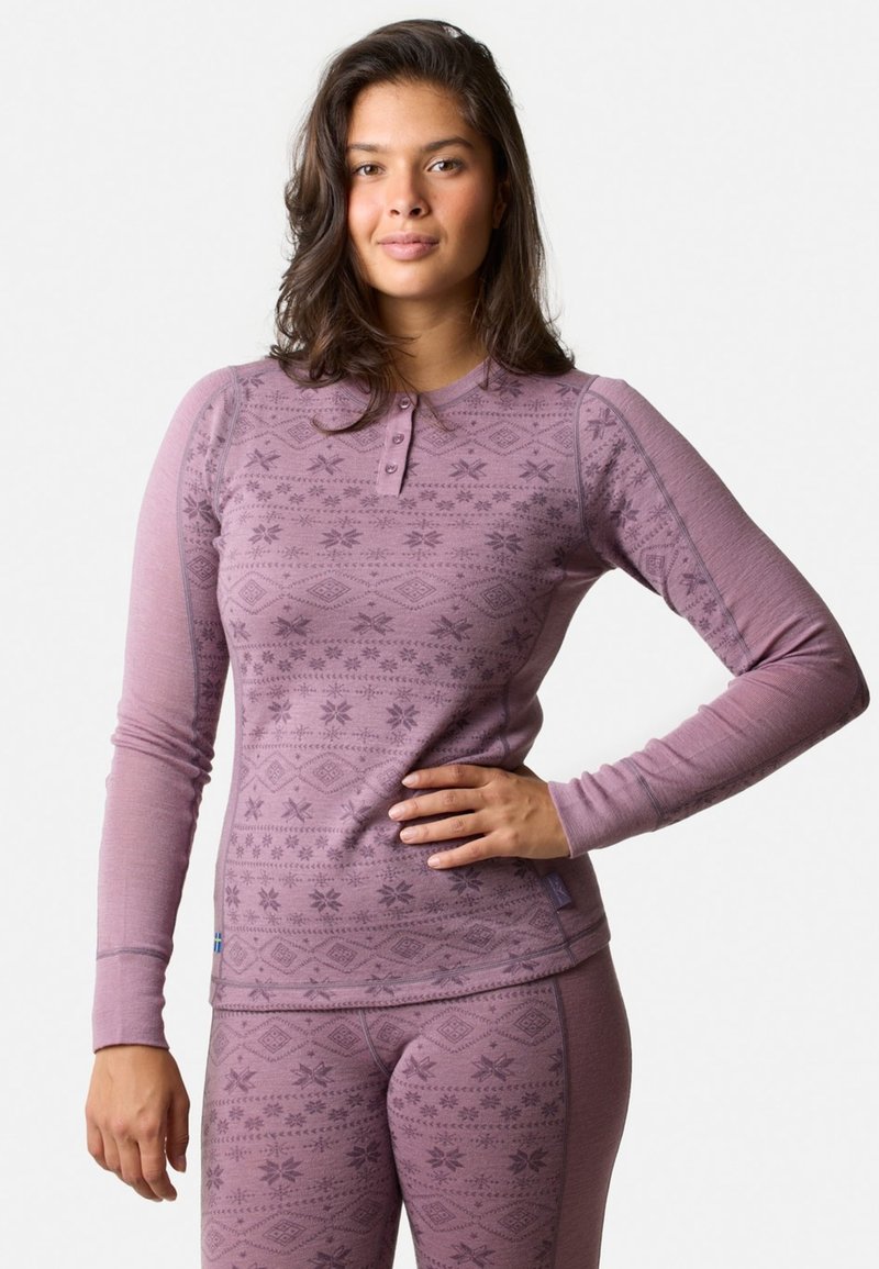 Ung kvinde iført matchende pink mønstrede termiske top og leggings, stående med den ene hånd på hoften mod en ensfarvet baggrund.