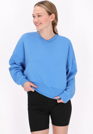 Lyseblå oversized sweatshirt med V-udskæring og ribbede kanter, parret med sorte tætsiddende shorts. Blød tekstur, afslappet pasform og lange ærmer.