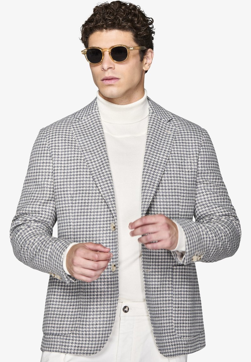 Grå houndstooth-blazer med to knapper, båret over en hvid turtle neck. Modellen bærer solbriller, hvilket fremhæver blazeren's tekstur og mønster.