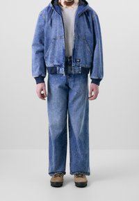 Džínová bunda s kapucí, šedé tričko, široké džínové kalhoty, béžové boty. Vlastnosti zahrnují zip a viditelné švy.