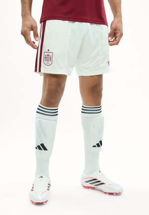 Joueur de football masculin portant un short blanc d'Espagne, des chaussettes blanches à rayures noires, des crampons blancs et un maillot rouge, debout sur fond blanc.