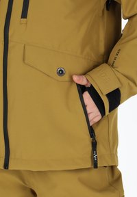 Braune Outdoor-Jacke mit einer Reißverschlusstasche, verstellbaren Bündchen und schwarzen Akzenten. Verfügt über ein wasserdichtes Material, das mit STX 20.000 gekennzeichnet ist.