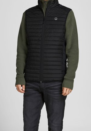 Homme portant une veste sans manches matelassée noire sur un pull en maille vert olive et un jean foncé, debout les mains détendues le long du corps.
