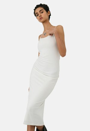 Fiatal nő rövid, göndör hajjal, szűk fehér ujjatlan midi ruhában és fekete csizmában, egyszerű háttér előtt pózol.