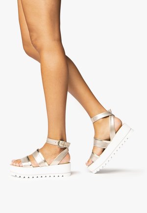 Sandales plates en métal doré avec plusieurs lanières et une sangle à boucle à la cheville. La semelle blanche présente un motif en zigzag pour une meilleure adhérence.