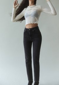 Top blanco transparente de manga larga y largo cropped con bordes en forma de ondas, combinado con jeans negros de tiro alto y botines negros, sobre un fondo claro.