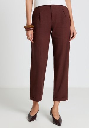 Frau trägt hoch taillierte, gerade braune Hose mit umgeschlagenen Säumen, kombiniert mit spitzen braunen Schuhen und klobigen Armbändern am linken Handgelenk.