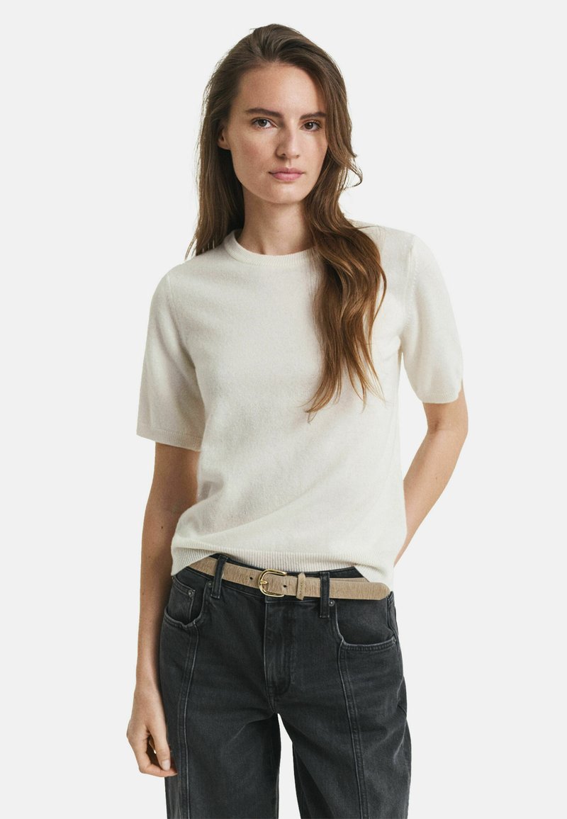 Cremfarbener Kurzarm-Strickpullover mit Rundhalsausschnitt, weicher Textur, kombiniert mit dunklen High-Waist-Jeans und einem hellen Gürtel.