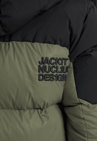 Black and olive green puffer jacket with black embroidered text reading "JACKIT NUCL1US DES1GN" on the olive section.
