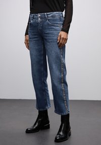 Vaqueros de mezclilla azul con un diseño de pierna recta, que presentan un sutil acento de estampado animal a lo largo del costado. Combinados con botas negras hasta el tobillo.
