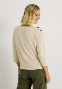 Pull tricot beige clair à manches trois quarts, avec de sombres boutons à l'épaule, et une coupe décontractée qui descend jusqu'à l'ourlet.