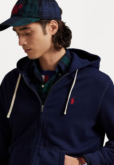 Tummansininen zip-up huppari, jossa on vaaleanbeiget nyörit ja pieni punainen logo. Sen alla näkyy vihreä ruudullinen paita, jossa on napitettava kaulus.