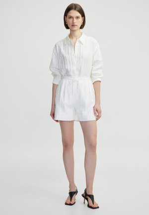 Mono de lino blanco con mangas largas, parte delantera con botones, tejido texturizado, cintura ajustable con cordón y dobladillo deshilachado; combinado con sandalias de tacón negro.