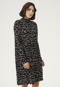 Robe noire avec un col montant, présentant un design plissé et un motif abstrait beige. Manches longues et coupe décontractée. Fabriquée en tissu léger.
