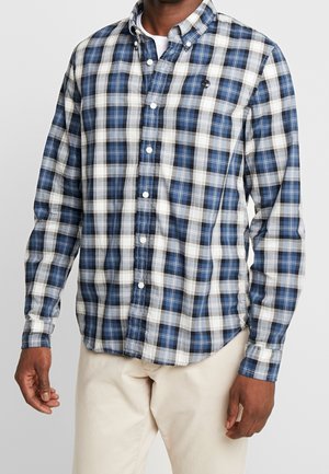 Mężczyzna w niebiesko-biało-czwartej koszuli na guziki z długimi rękawami i beżowych spodniach, stojący z rękami swobodnie opadającymi wzdłuż ciała.